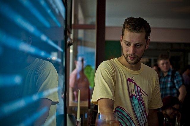 A man standing beside a window that reflects blue screen light, in a busy cafe
